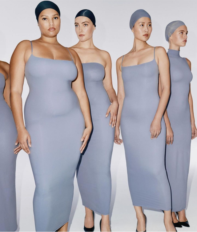 Five people stand together wearing various different styles of a long slip dress in a slate grey colour, they are also wearing caps.