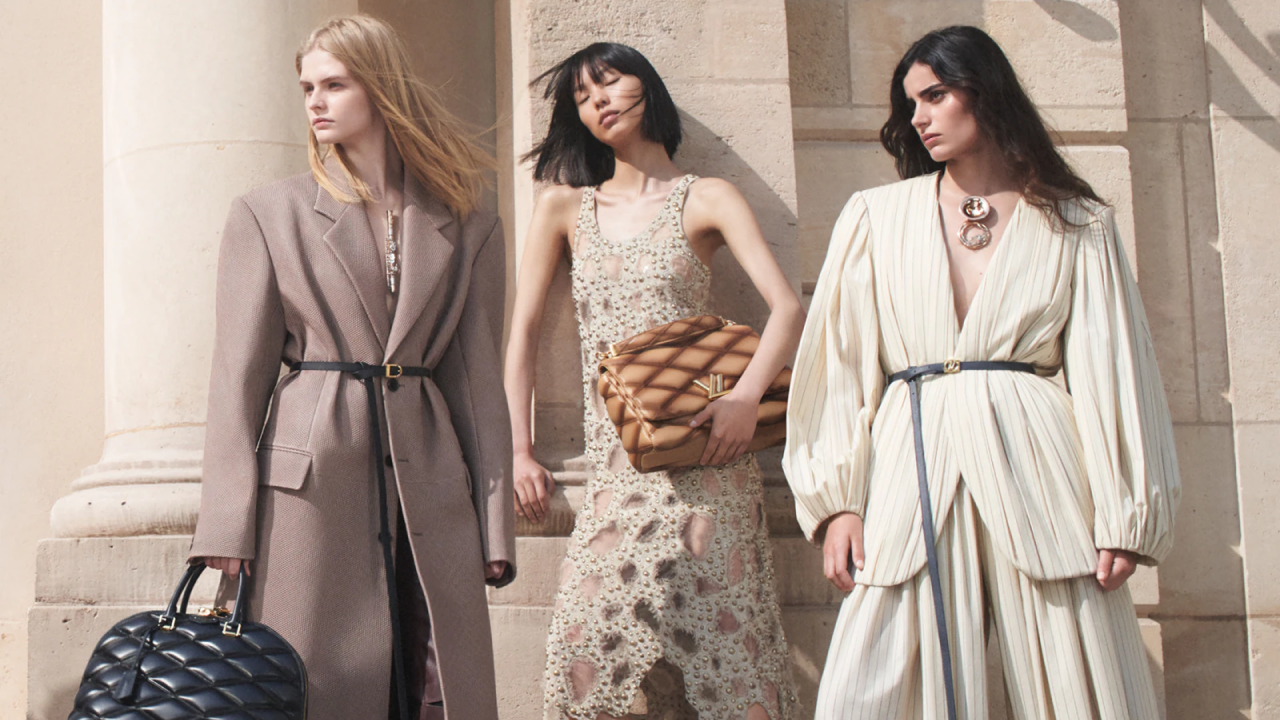 Three people stand against pillars. The first wears a long taupe coat and a large quilted black handbag. The second wears a cream, studded, long evening dress covered in holes and an oversized, brown, quilted clutch handbag. The third wears a pin stripped, long sleeved jacket style top and trousers.