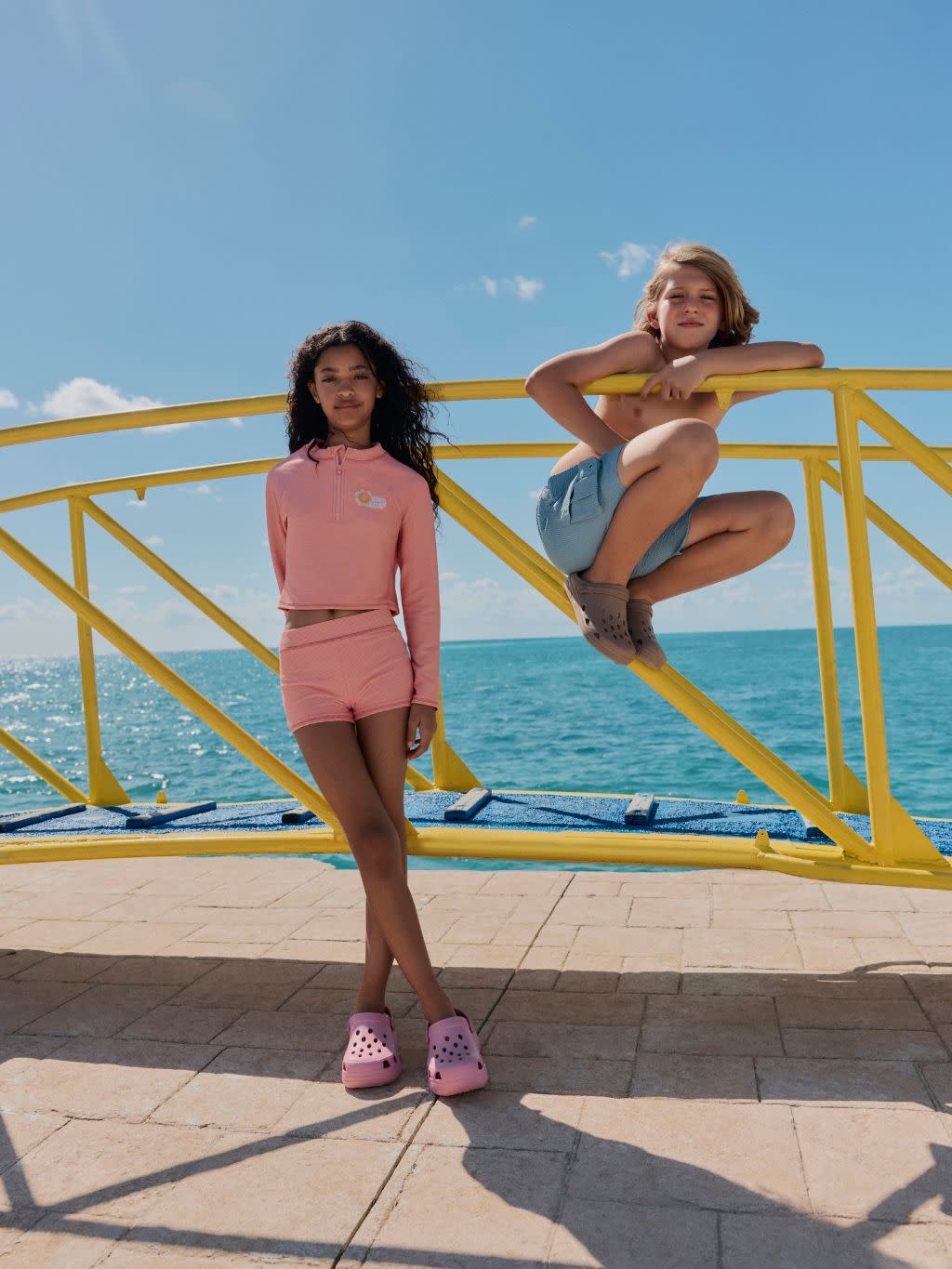 A boy and a girl wearing pastel swimwear and crocs standing on a seafront