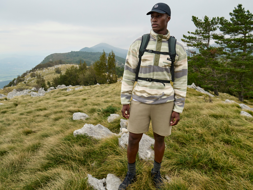 A man stands on a hill dressed in a fleece and cargo shorts with a rucksack on his back